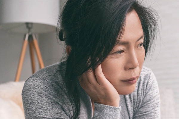 A middle-aged asian woman sitting on her couch, resting her head on her hand, suffering from adrenal fatigue. Get treatment for adrenal fatigue syndrome from Tasmin Cordie, D.C. of Sage Functional Medicine in Marlboro Township.