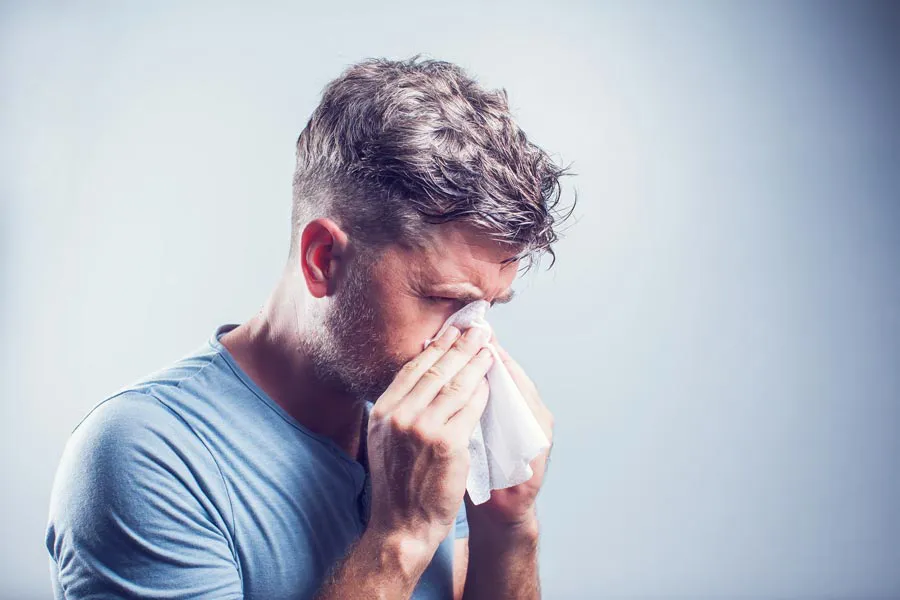 A man in a gray shirt blows his nose. Get help for allergies from Tasmin Cordie, D.C. of Sage Functional Medicine in Marlboro Township.
