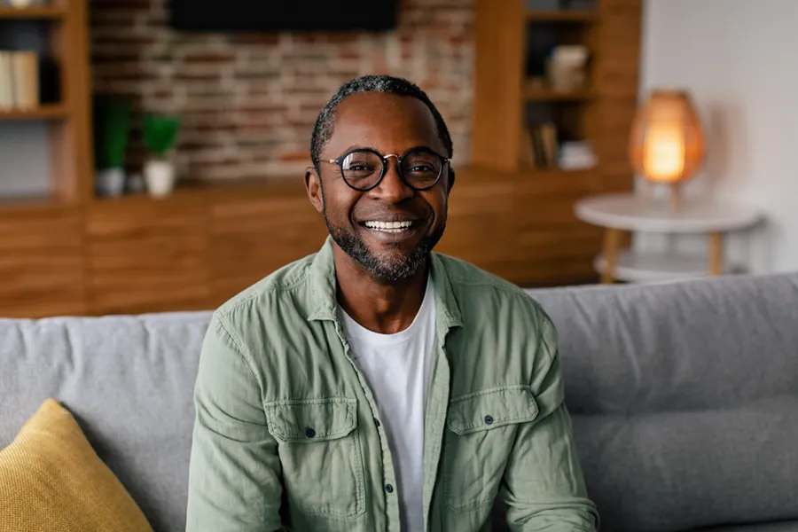 A middle aged black man in a green button up shirt sits on his couch smiling, happy with his treatment for andropause and low testosterone from Tasmin Cordie, D.C. of Sage Functional Medicine in Marlboro Township.