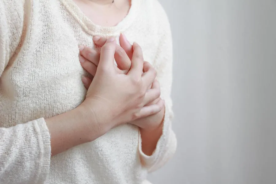 Close-up of a woman in a white sweater clutching her chest in respiratory distress. Get treatment with asthma from Tasmin Cordie, D.C. of Sage Functional Medicine in Marlboro Township.