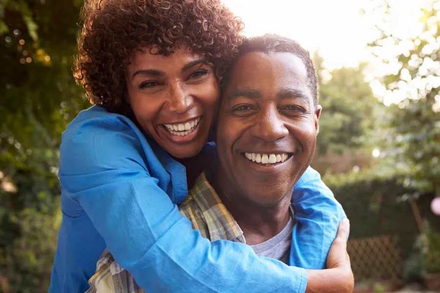 A middle-aged African American couple outside, the woman embracing the man from behind pressing her cheek to his, both are smiling due to successful Autoimmune Disorder treatment from Tasmin Cordie, D.C. of Sage Functional Medicine in Marlboro Township.