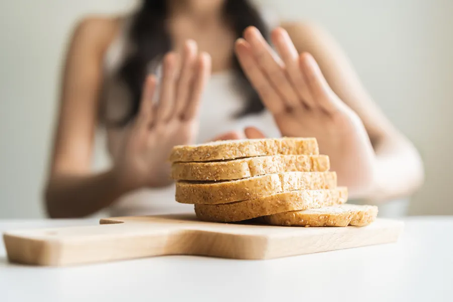 Close up of a stack of bread slices on a cutting board with a woman's hands behind them, refusing to eat. Get treatment for Celiac Disease from Tasmin Cordie, D.C. of Sage Functional Medicine in Marlboro Township.