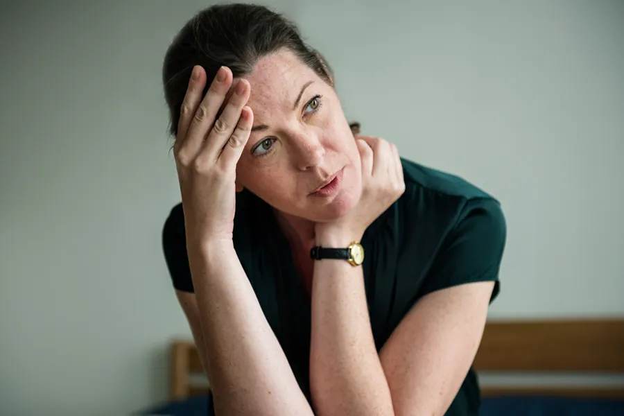 A woman in a dark blue dress sits on her bed holding her head in her hands with an exhausted expression. Get help for chronic fatigue syndrome from Tasmin Cordie, D.C. of Sage Functional Medicine in [AERA].