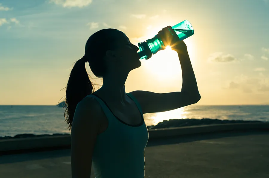 A woman drinks from a bottle of water during sunset at the beach. Get treatment for detoxification challenges from Tasmin Cordie, D.C. of Sage Functional Medicine in Marlboro Township.