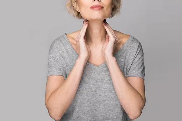 An older blonde woman in a gray t-shirt holds her hands to her neck checking for signs of thyroid problems. Get help for Hashimoto's Thyroiditis from Tasmin Cordie, D.C. of Sage Functional Medicine in Marlboro Township.