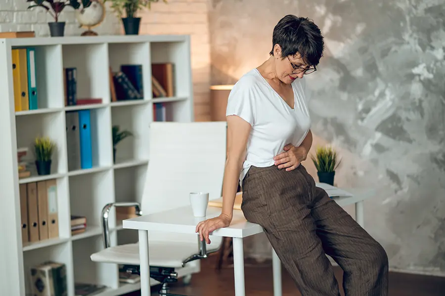 A woman leans against her desk and holds her stomach. Schedule treatment for irregular digestion from Tasmin Cordie, D.C. of Sage Functional Medicine in Marlboro Township.