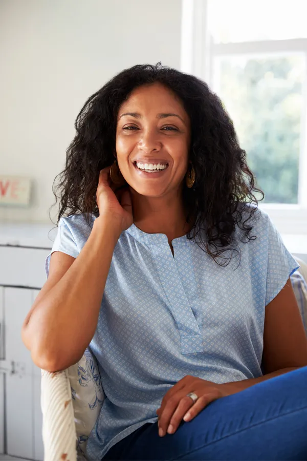 A middle-aged Black woman sits and smiles in her living room. Get treatment for irregular digestion from Tasmin Cordie, D.C. of Sage Functional Medicine in Marlboro Township.