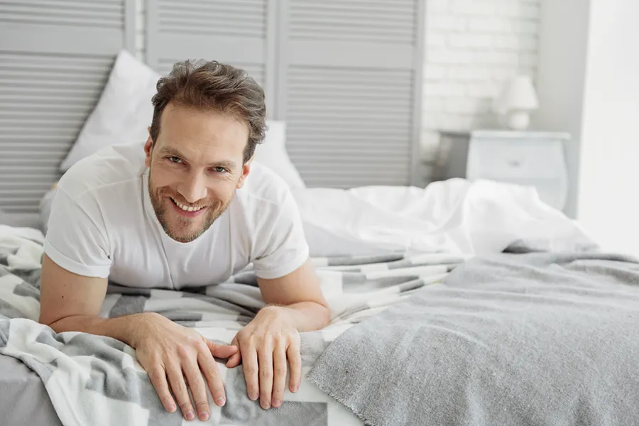 A dirty-blonde haired man lays on his stomach in bed smiling. Schedule treatment for low libido from Tasmin Cordie, D.C. of Sage Functional Medicine in Marlboro Township.