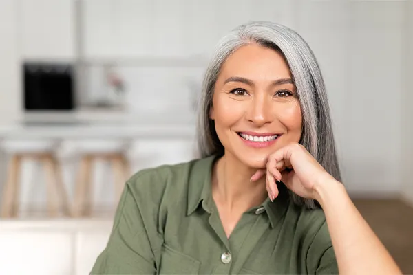 An asian woman with long gray hair and a green shirt sitting and smiling on her couch, happy with her menopause care from [NAME} in Marlboro Township.