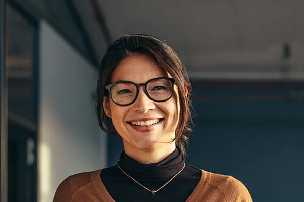A brunette woman with glasses smiles in a gray-blue walled office. Get a comprehensive consultation for metabolic imbalance from Tasmin Cordie, D.C. of Sage Functional Medicine in Marlboro Township.