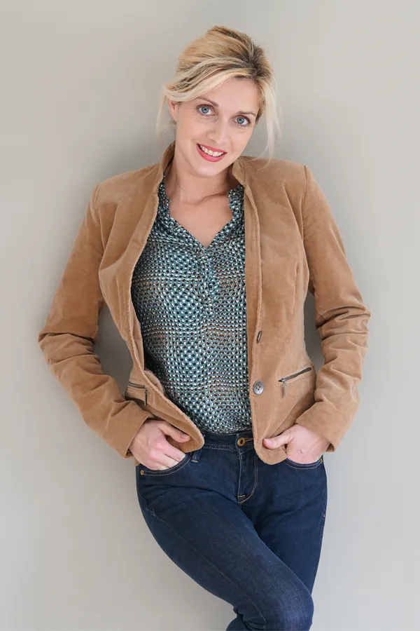 A smiling blonde woman in a tan leather jacket leans against a gray wall. Schedule metabolic imbalance consultation from Tasmin Cordie, D.C. of Sage Functional Medicine in Marlboro Township.