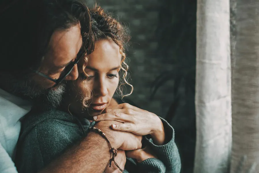 A man embraces a woman from behind in a dimly lit room, their somber expressions showing concerns about a neurodegenerative disease they will get treated by Tasmin Cordie, D.C. of Sage Functional Medicine in Marlboro Township.