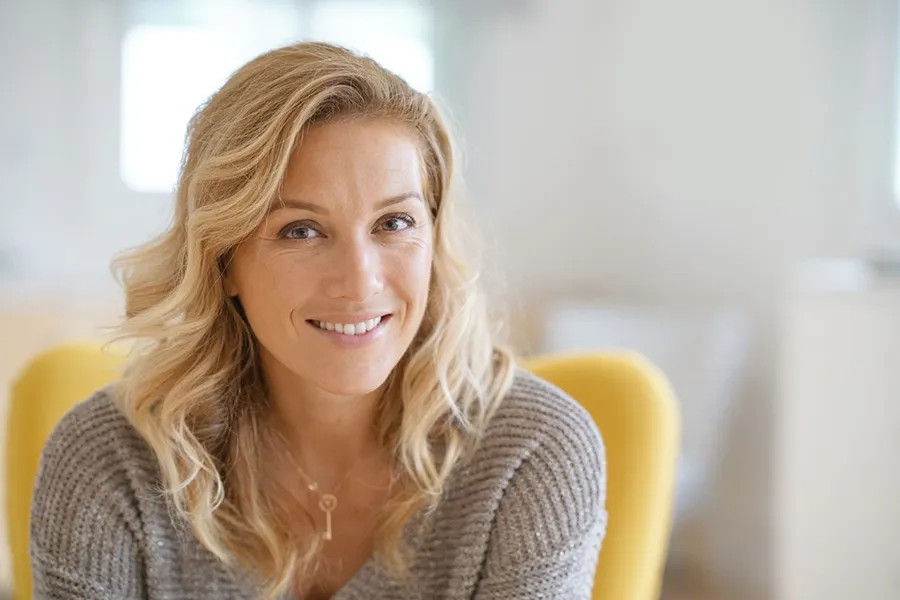 A blonde woman wearing a a gray sweater sits inside in a yellow chair, smiling and happy with her perimenopause treatment from Tasmin Cordie, D.C. of Sage Functional Medicine in Marlboro Township.